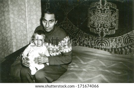 UKRAINE, ZAPOROZHYE REGION, VILLAGE SAMOYLOVKA - 1978: A vintage photo shows man sitting on a bed and holding a little girl. Name of girl id  is Julia Voronkov