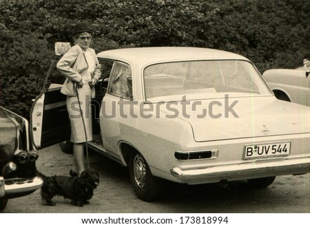 GERMANY, WEST BERLIN - 1950s: An antique photo shows woman in elegant suit and hat with a dog on a leash, straightens her skirt at the parking near the open door of Opel