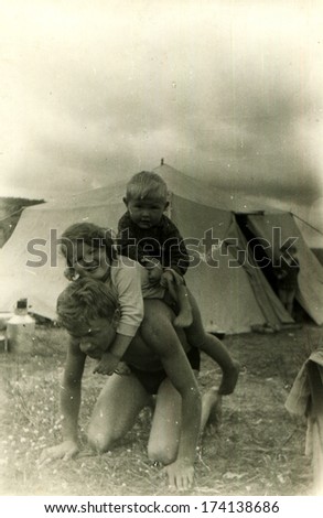 GERMANY -  1950s: An antique photo shows little girl and boy sitting on the back of his father, who was kneeling on the background of the tent