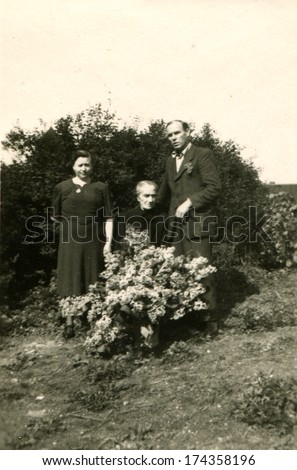 GERMANY, ROSTOCK -  CIRCA 1930s: An antique photo shows Man and woman posing next to an old woman sitting with a bouquet of flowers on a background of bushes