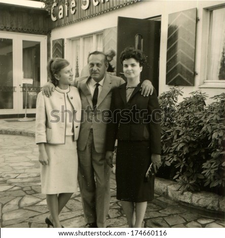 GERMANY -  CIRCA 1960s: An antique photo shows man in a business suit hugging the shoulders of two elegant women in front of the cafe