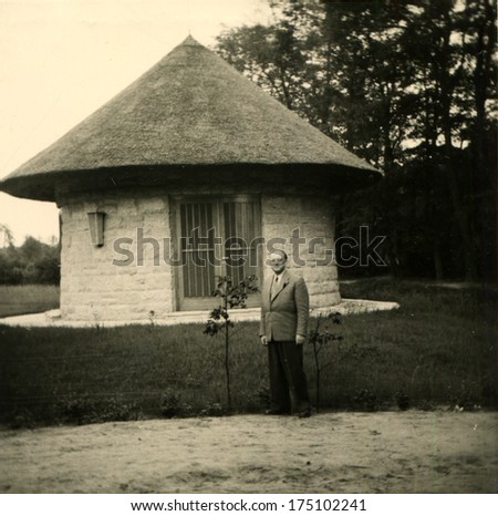 GERMANY -  CIRCA 1959: An antique photo shows man posing against the backdrop of one-floor round house