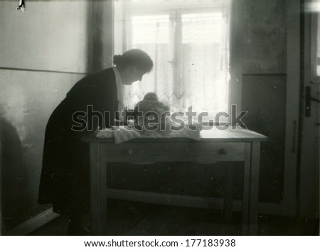 GERMANY - CIRCA 1937: An antique photo of middle-aged woman leaned over the table near the window, on which lies the infant