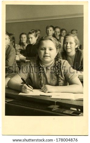 GERMANY - January 8, 1956: An antique photo of full-faced schoolgirl with plaits sits at his desk with a pen in hand on background of classmates