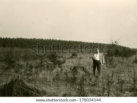 GERMANY -  CIRCA 1970s: An antique photo shows man in a jacket thrown over his shoulders, posing in front of overgrown fur trees aganst  forest