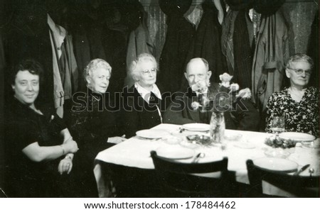 GERMANY - CIRCA 1950s: An antique photo of group of elderly people sitting at a table against the background of hangers with outerwear