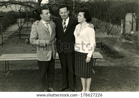 GERMANY - CIRCA 1960s: An antique photo of middle-aged man embracing shoulders wife and adult son, men in suits and ties, all three are in the garden