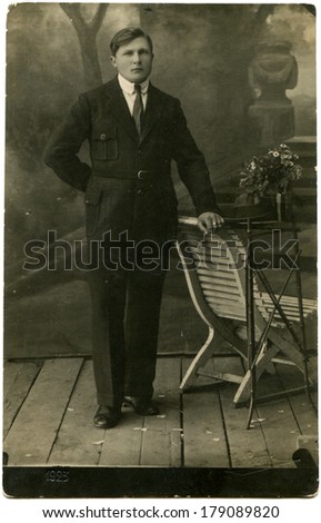 ESTONIA, VALGA - CIRCA 1925: A vintage photo of studio portrait of a young man in a business suit posing beside the table with flowers and a wooden chair