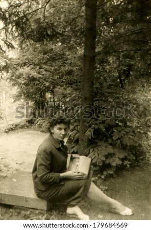 GERMANY - CIRCA 1950s: An antique photo of party woman with a bag on her lap sitting on a stone slab under spruce