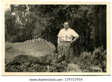 GERMANY - CIRCA 1950s: An antique photo of mature man in a white shirt with short sleeves standing near a large boulder on the background of trees