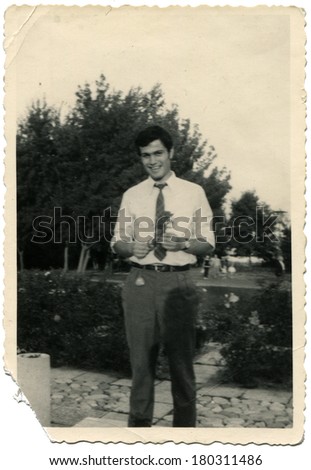 GERMANY - CIRCA 1950s: An antique photo of young man in white shirt and tie, with a rose in his hands, posing on a background of park