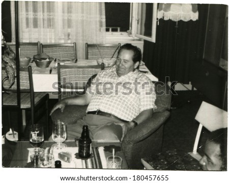 GERMANY, BERLIN - CIRCA 1960s: An antique photo of middle-aged man sitting in a armchair in front of a coffee table on which stand the bottle and a few glasses of beer, Berlin