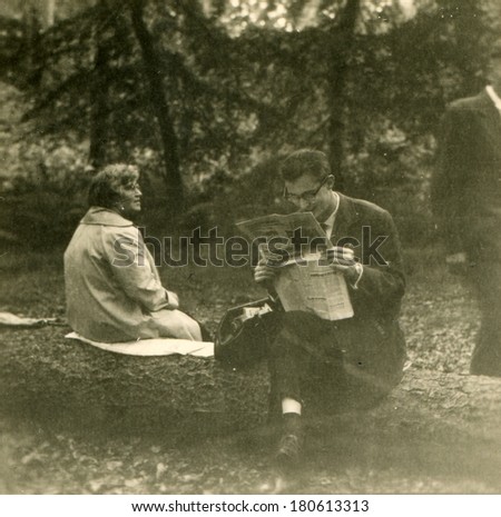 GERMANY, ROSTOCK - CIRCA 1950s: An antique photo of young man in a business suit reading a newspaper while sitting on a log in the park, next to him with his back to the camera sits middle-aged woman
