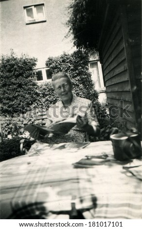 GERMANY, BAD DOBERAN - CIRCA June, 1960: An antique photo of woman reading newspaper at a table in garden near the wall of wooden plank house, Bad Doberan, district of Rostock, Mecklenburg-Vorpommern