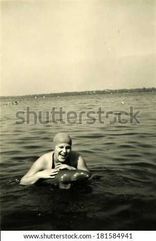 GERMANY - CIRCA 1950s: An antique photo of laughing young woman in rubber bathing cap bathing sea with inflatable toy