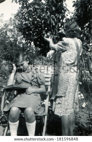 GERMANY - CIRCA 1950s: An antique photo of girl reading a book while sitting in a folding chair in the garden, next to her is a woman and tear apple from a tree