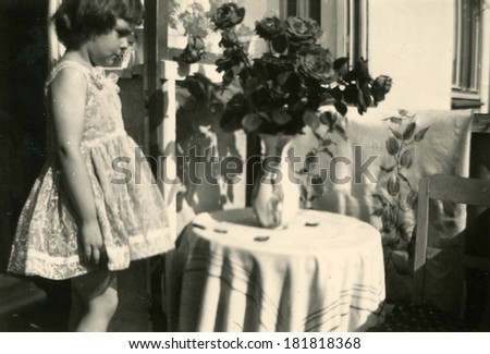 GERMANY - CIRCA 1940s: An antique photo of girl in a celebratory dress stands near the round table covered with a tablecloth, on which is a vase with a bouquet of roses, near the home