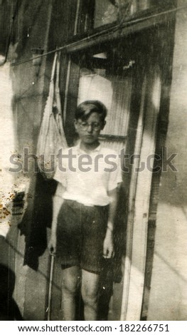 GERMANY, POTSDAM - CIRCA 1930s: An antique photo of boy with glasses, white shirt with short sleeves and shorts posing doorway, Potsdam