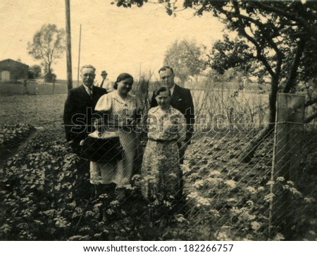 GERMANY, ROSTOCK - CIRCA 1930s: An antique photo of two men, two women and a little boy with accordion posing in the garden behind the fence of the grid-netting
