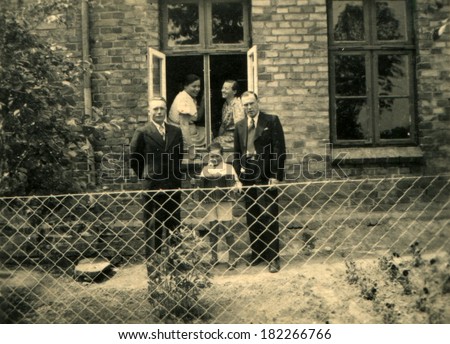 GERMANY, ROSTOCK - CIRCA 1930s: Antique photo of Two men and little boy with accordion posing behind fence from the grid-netting on the background of a brick house in the window of which sit two women