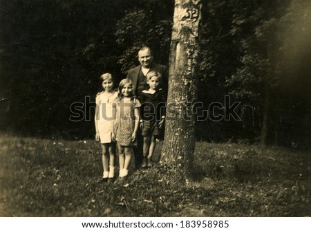 GERMANY - CIRCA 1940s: An antique photo of full middle-aged man in a three-piece suit posing with two girls and a boy near a tree in a clearing in a forest