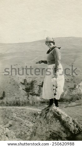 CANADA - CIRCA 1950s: Reproduction of an antique photo shows woman in a white skirt and hat standing on a rock on a background of mountains