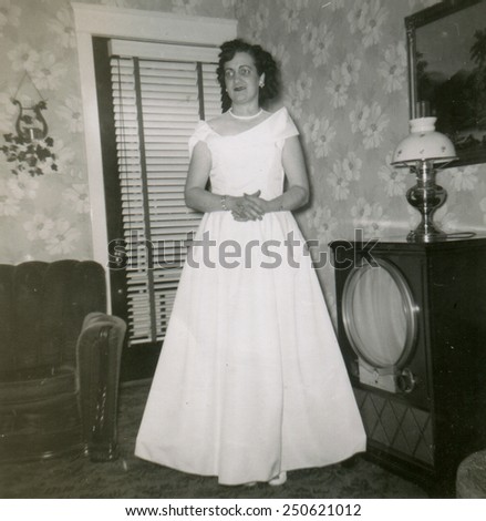 CANADA - CIRCA 1950s: Reproduction of an antique photo shows mature woman in a white evening dress posing in the living room near a radio