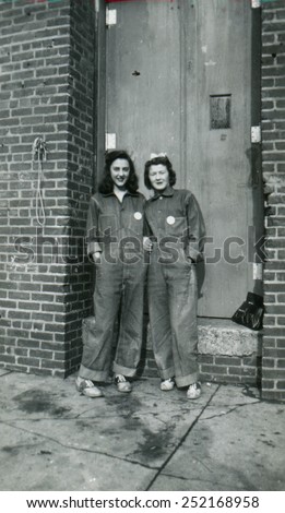 CANADA - CIRCA 1940s: Reproduction of an antique photo shows Two girls in denim overalls standing near the door of a brick industrial building