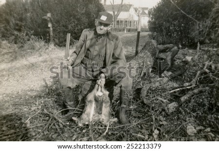 CANADA - CIRCA 1940s: Reproduction of an antique photo shows hunter with a gun sitting on a felled tree on background of dirt road leading to private home, and holds in his hand the three killed hares