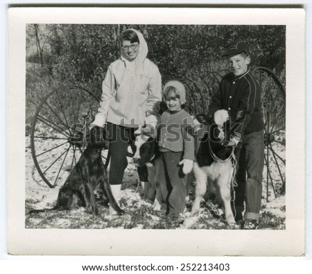 CANADA - CIRCA 1940s: Reproduction of an antique photo shows young woman in a white jacket and glasses posing with warmly dressed boy and girl with small bulls and a large black dog