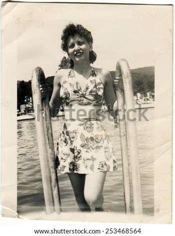 USA - CIRCA 1944: Reproduction of an antique photo shows young woman in swimsuit posing on the ladder of the ship on a background of water and shore resort