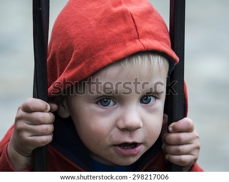 Little boy in a red jacket with a hood looks out for wrought fence