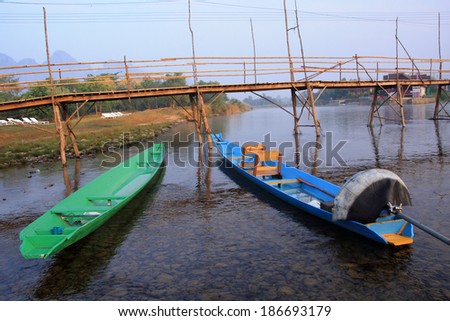 a blue boat and a green boat float side by side in a small river under a wooden bridge