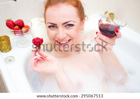 Young woman with strawberry and wine while lying in the bath with foam and candles