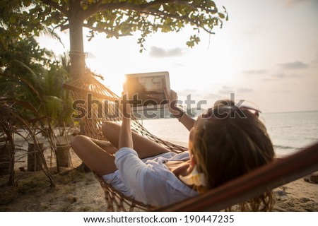 Woman enjoying sunset view by the beach on hammock using digital tablet