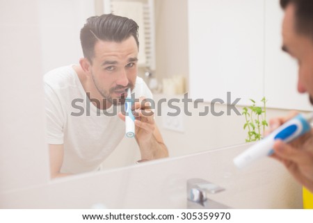 Joyful man looking at camera in mirror reflection during brushing teeth with an electric toothbrush