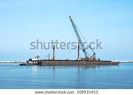 A tugboat pushes a floating crane on a barge and other repair equipment and materials toward the breakwall between the open waters of Lake Erie and the harbor at Cleveland, Ohio