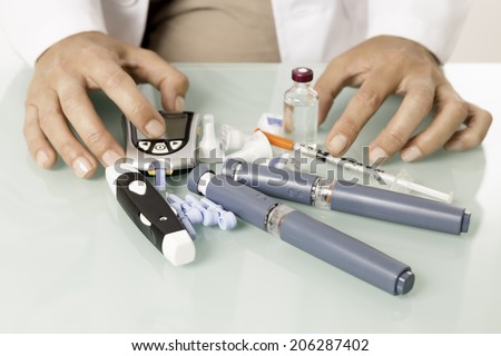 closeup of hands of a doctor presenting vial of insulin, glucometer, lancets, syringe, reactive strips, insulin pens and a lancing over the desk - focus on insulin pens