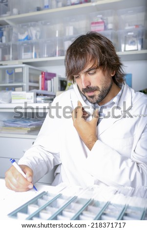 young male pharmacist talking on the phone at pharmacy sitting in front of a desk with pillboxes taking an order on a notebook - focus on the pharmacist