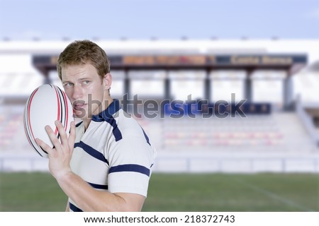 portrait of a young male rugby player standing holding a rugby ball in his hand on a rugby field background