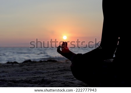 backlight of a woman practicing yoga sitting on the shore of the beach at sunset simulating holding the sun in her finger - focus on the fingers