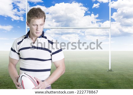 portrait of a young male rugby player standing holding a rugby ball in his hand on a green rugby field background