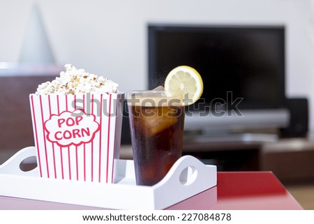 closeup of a classic red and white paper box of popcorn and a glass with cola, ice cubes and a slice of lemon on a table in a sitting room and a lcd television at background - focus on the box