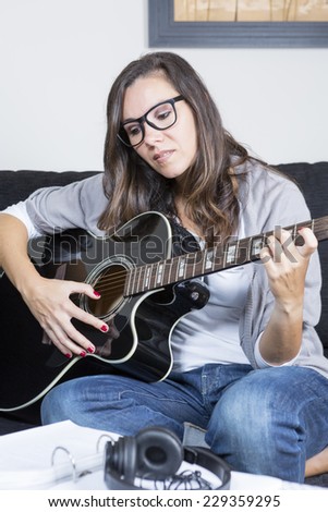 young woman playing guitar sitting on a sofa at home with headphones and sheet music book