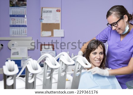 young female dental clinic assistant smiling is putting the disposable dental bib to a female patient sitting on a dental chair at the cabinet