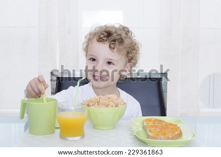 happy child is enjoying breakfast sitting on a chair, consists of a glass of natural orange juice, a bowl of cereals, a cup of milk and a toast with butter and peach mermelade