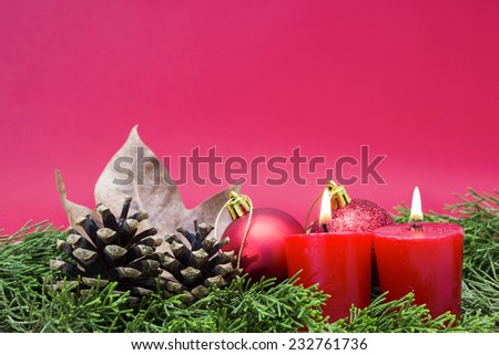 Christmas composition consisting of two red candles lighted, two shiny red balls, pinecones and dry leaves on fir branches on a red background