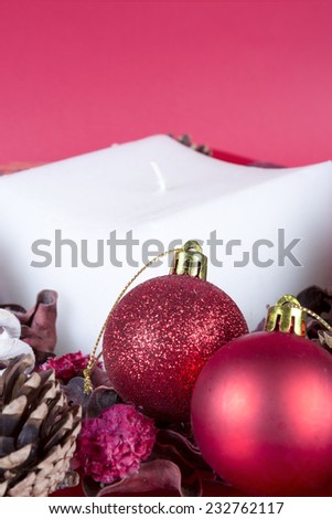 closeup of a Christmas composition consisting of a square white candle, two shiny red balls, pine cones and dry leaves on a red background - focus on the red ball