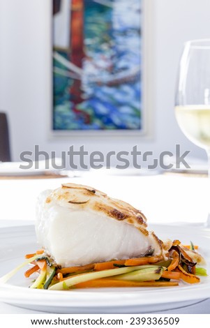 closeup of a codfish loin baked with grilled vegetables served on a plate and a glass of white wine on a restaurant background