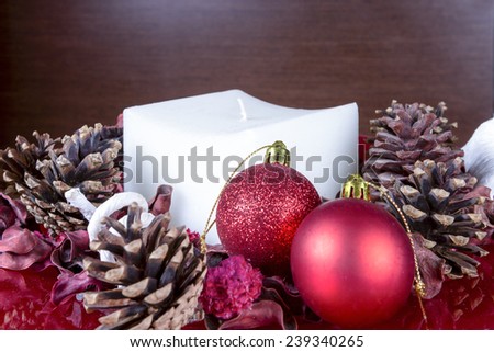 closeup of a Christmas composition consisting of two shiny red balls, a square white candle, pinecones and dry leaves on a red base on a wood background - focus on the red ball